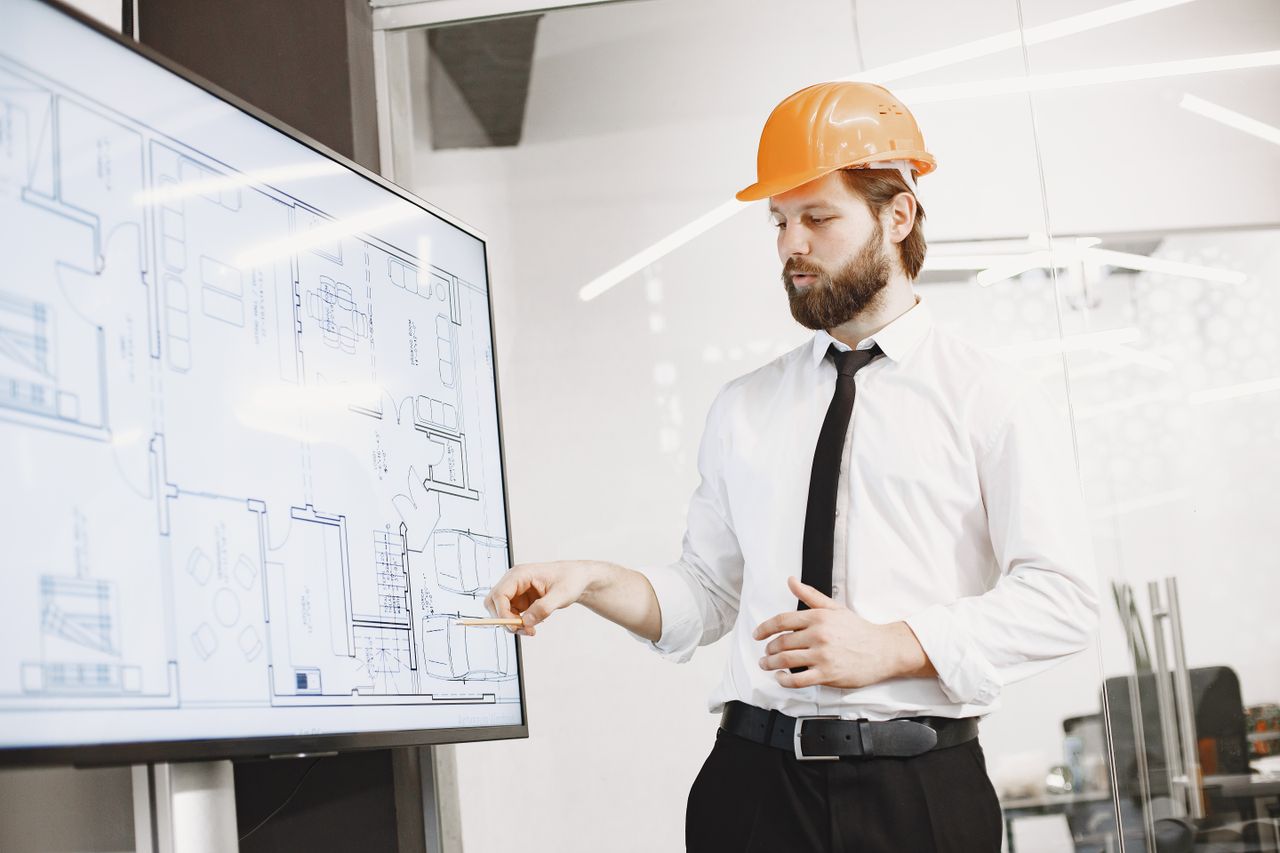 creative-03 Architect in hard hat reviewing blueprints on a screen, discussing plans in modern office.